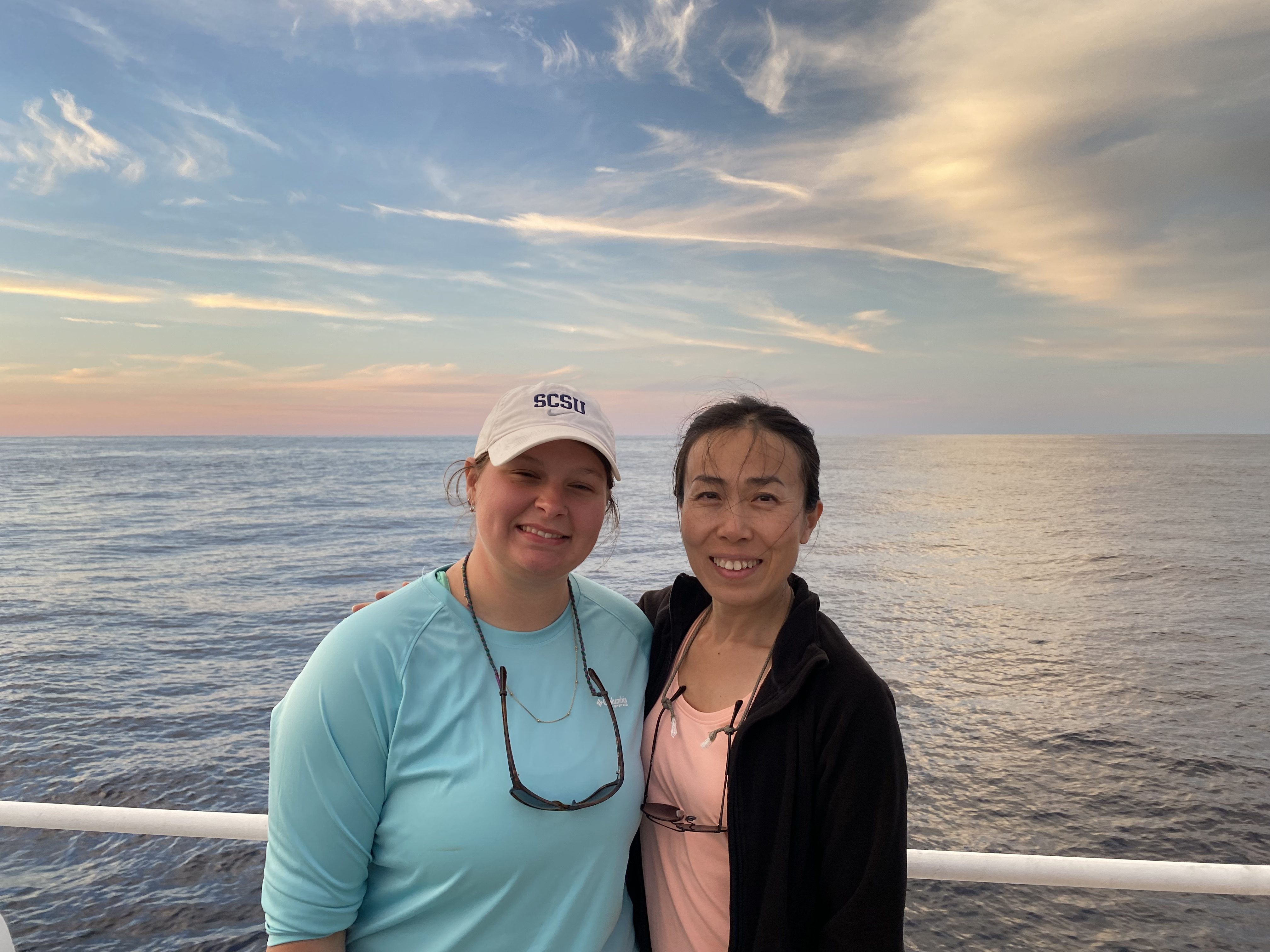 Two women standing side by side smiling at the camera while on a boat in the middle of an ocean, the sky is blue and pink.