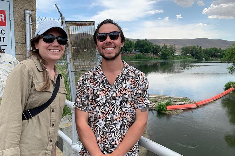 Two smiling people stand on a structure in front of a bridge