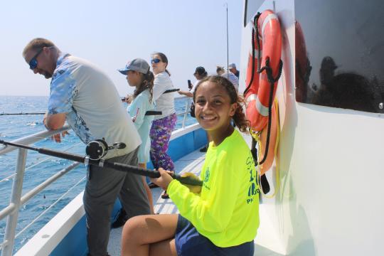 A young girl in pigtails holds a fishing rod and smiles at the camera. She it sitting on a white boat with other people fishing along the side of the boat behind her.