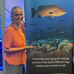 Woman stands besides and gestures to a tall, big blue banner with a photograph of fish and coral reefs for the Flower Garden Banks National Marine Sanctuary.
