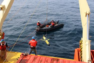 Researchers in hard hats and vests use a pulley system to lower a glider into the water off the back of a research ship. Other researchers sit in a black raft floating in the water behind the boat.